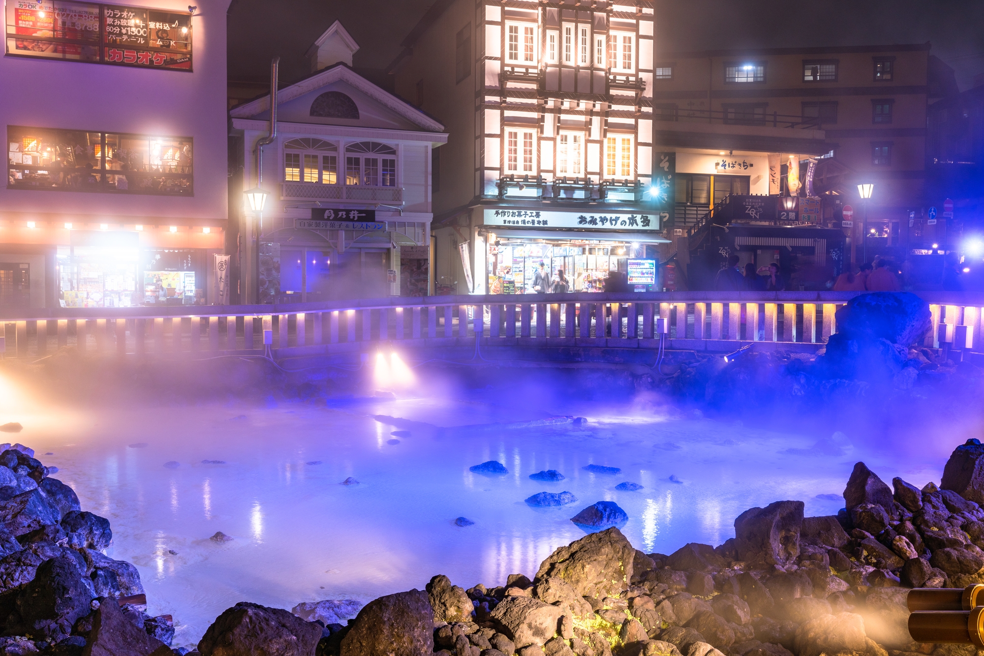 An outdoor hot spring at night, in front of a brightly illuminated hotel building