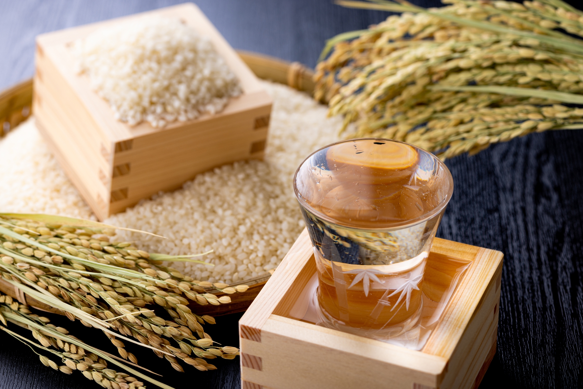 Sake in a small glass, overflowing into a wooden box. Polished rice and rice stalks are in the background