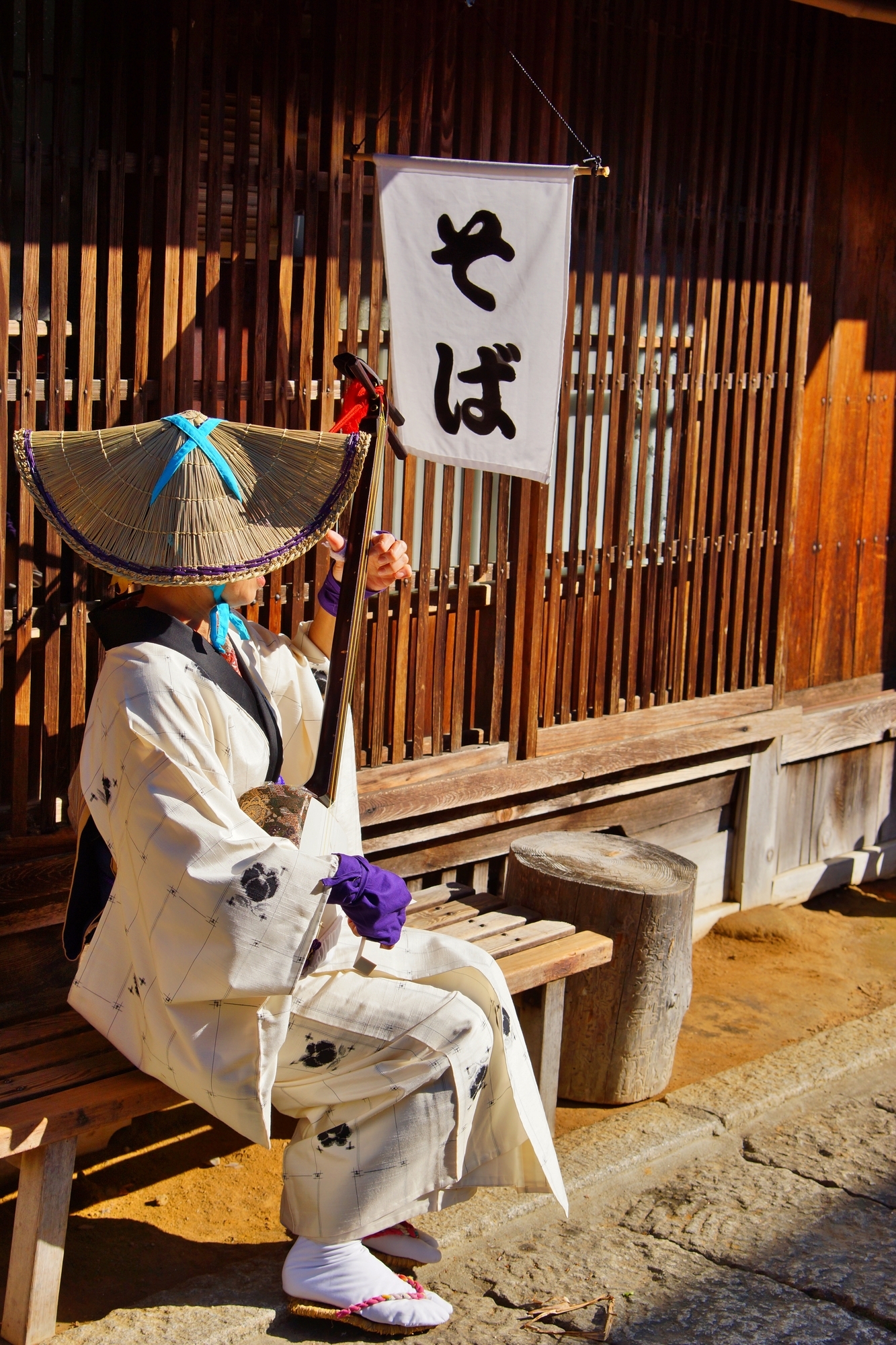 A woman in Edo-era kimono and straw hat playing an instrument outside a restaurant