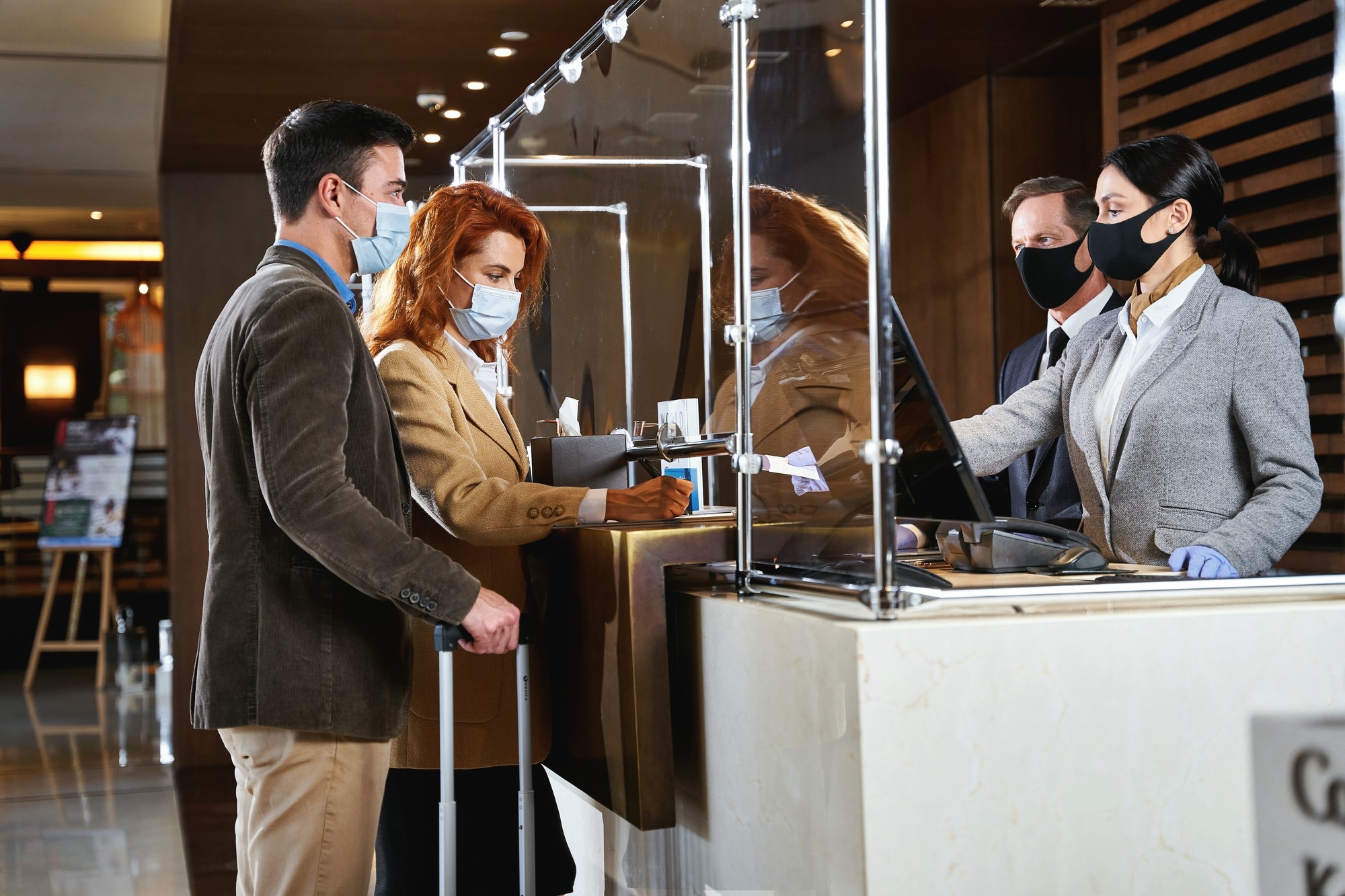 A man and a woman check in at a hotel, guests and employees are all wearing masks